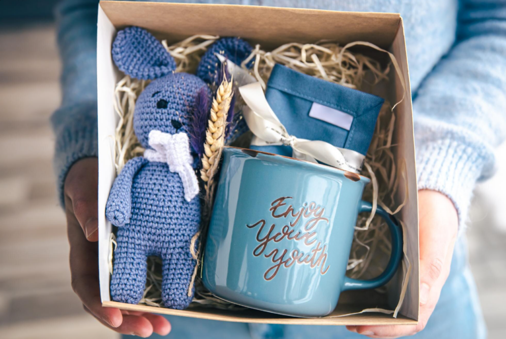 Offenes Geschenk, gehalten von einer Person in blauem Kleid, nur Hände sichtbar. Inhalt: gehäkelter blauer Hase, blaue Tasse und blaues Etui.